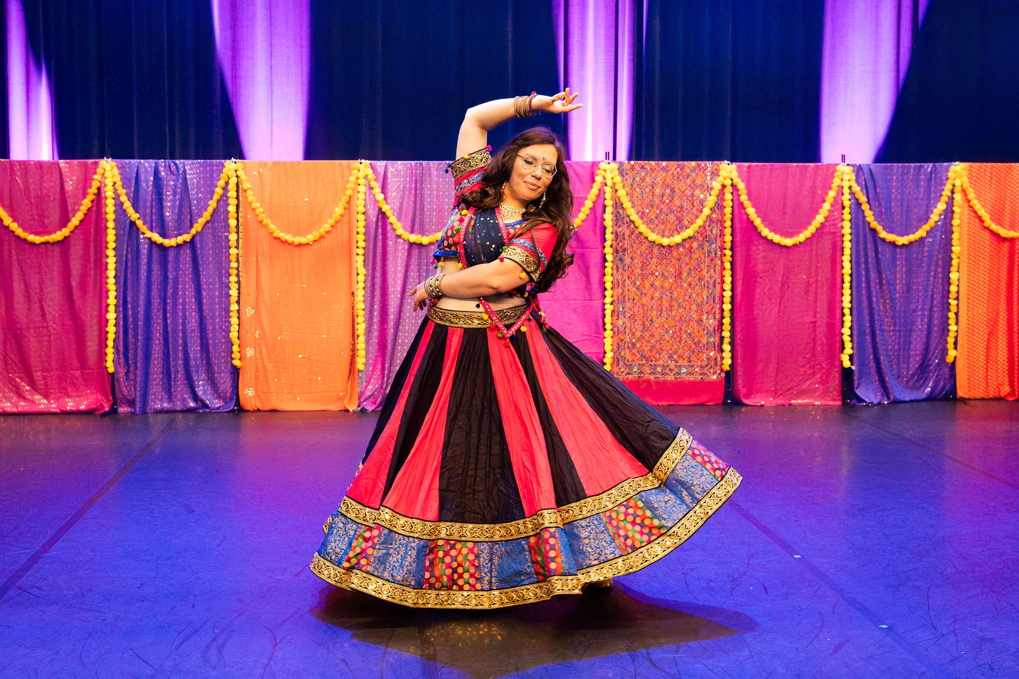 Un femme en costume de danse indienne pose sur scene avec un bras au dessus de sa tete et un bras faisant le tour de sa taille. Sa jupe tourne en eventail.