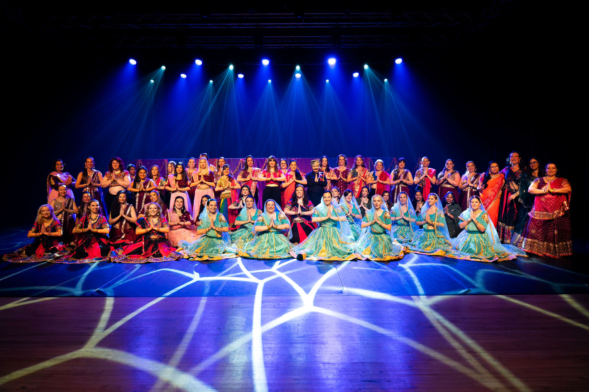Un groupe de plus de 50 danseuses en tenue indienne ou bollywood pose sur scene. 